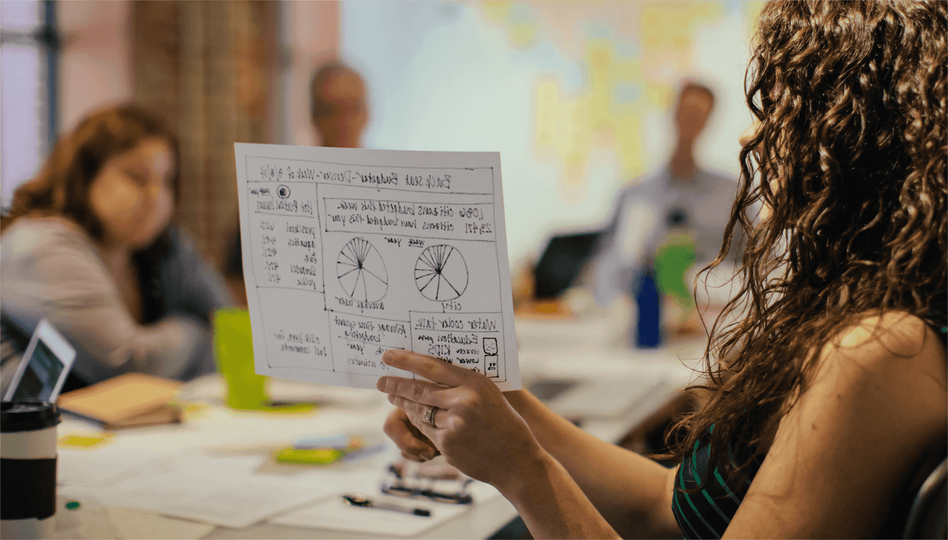 A woman hold a hand-drawn wireframe sketch of the Balancing Act platform while describing the features during a design workshop