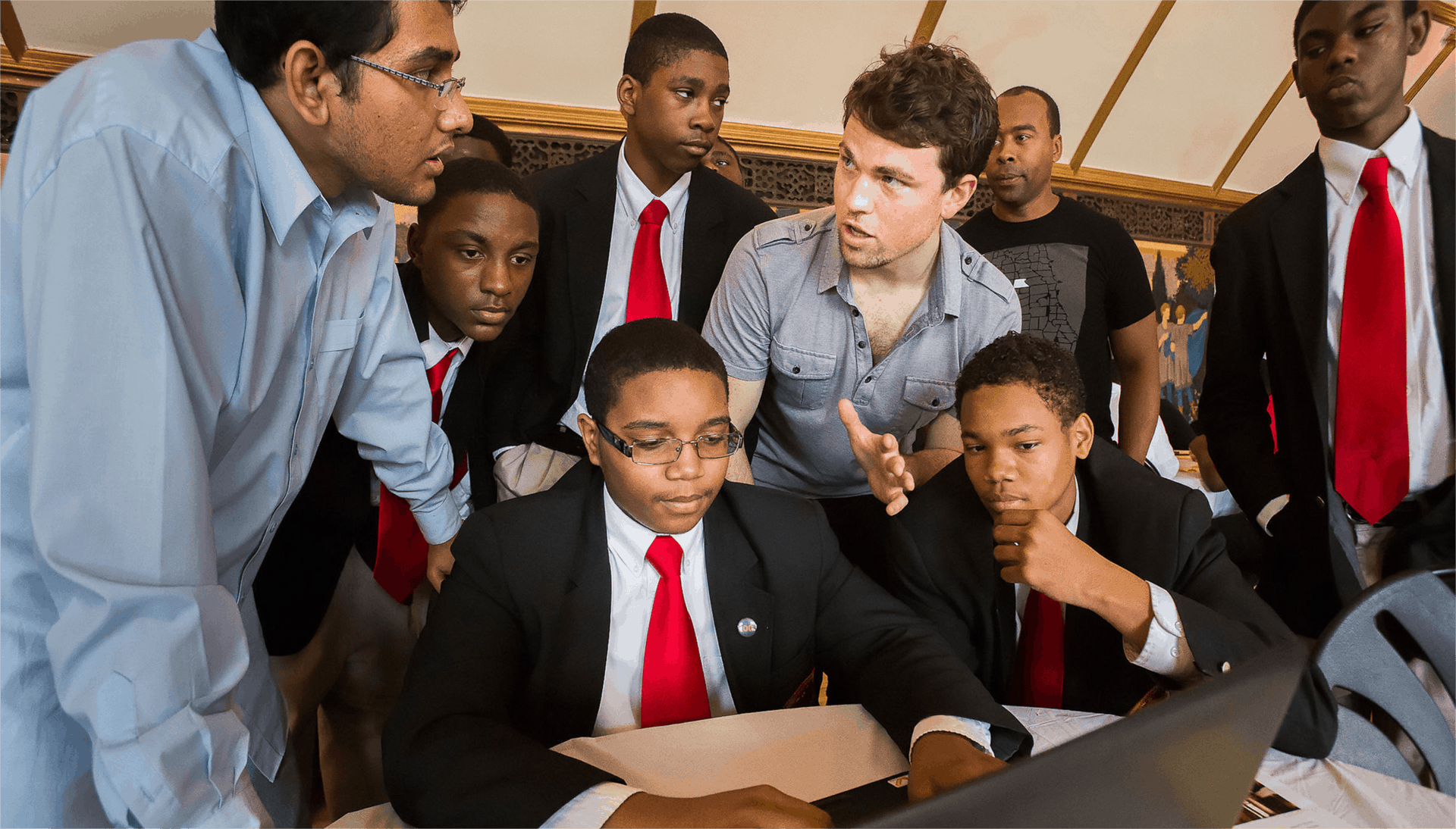 A group of students and teachers sit gathered around a laptop during a design workshop