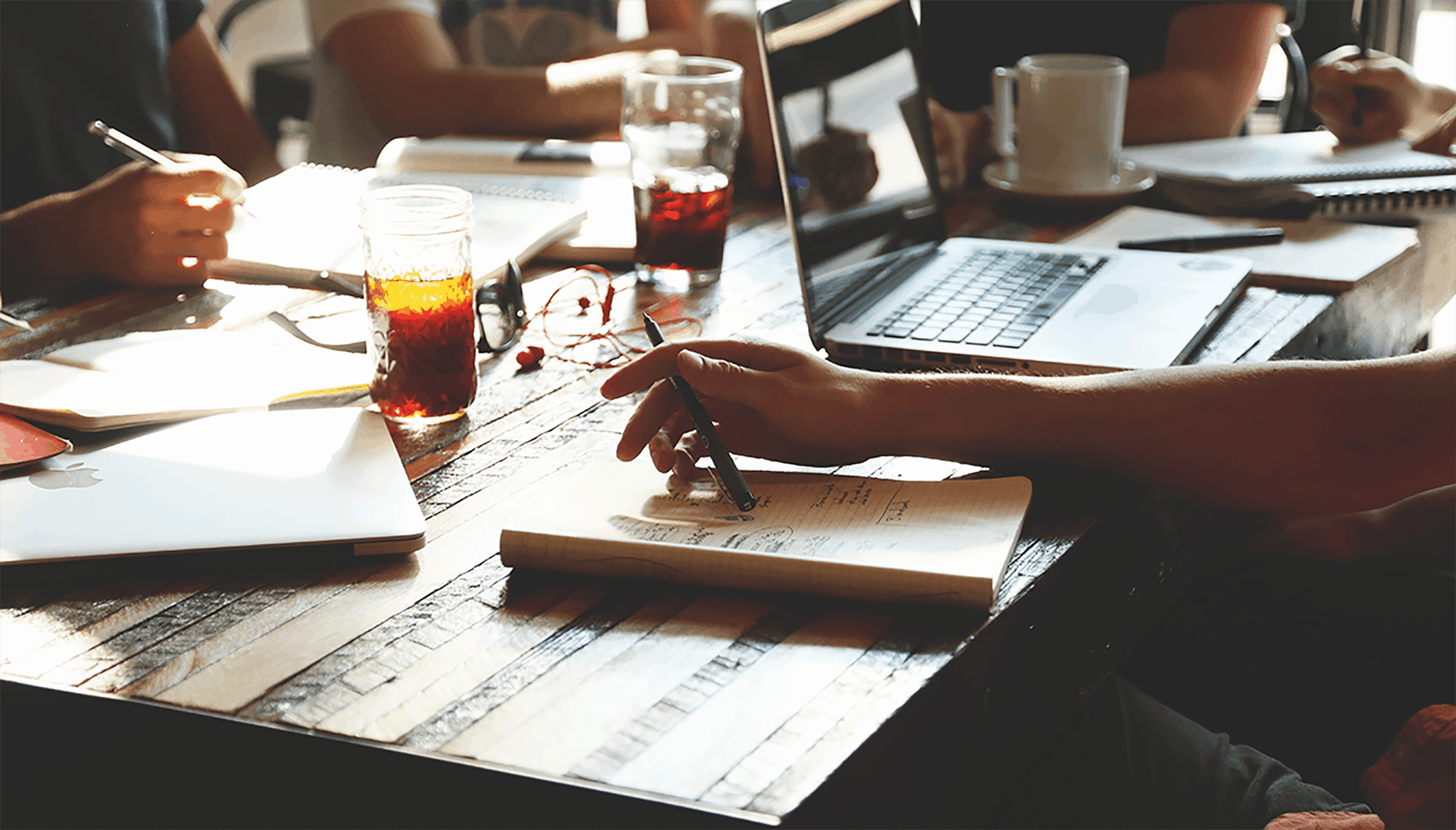 A group of people work in notebooks at a table lit by sunlight.