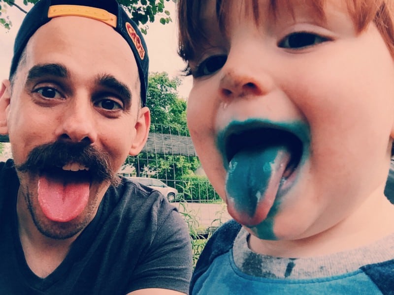 A man and child stick their tongues out. The child's tongue is blue from candy