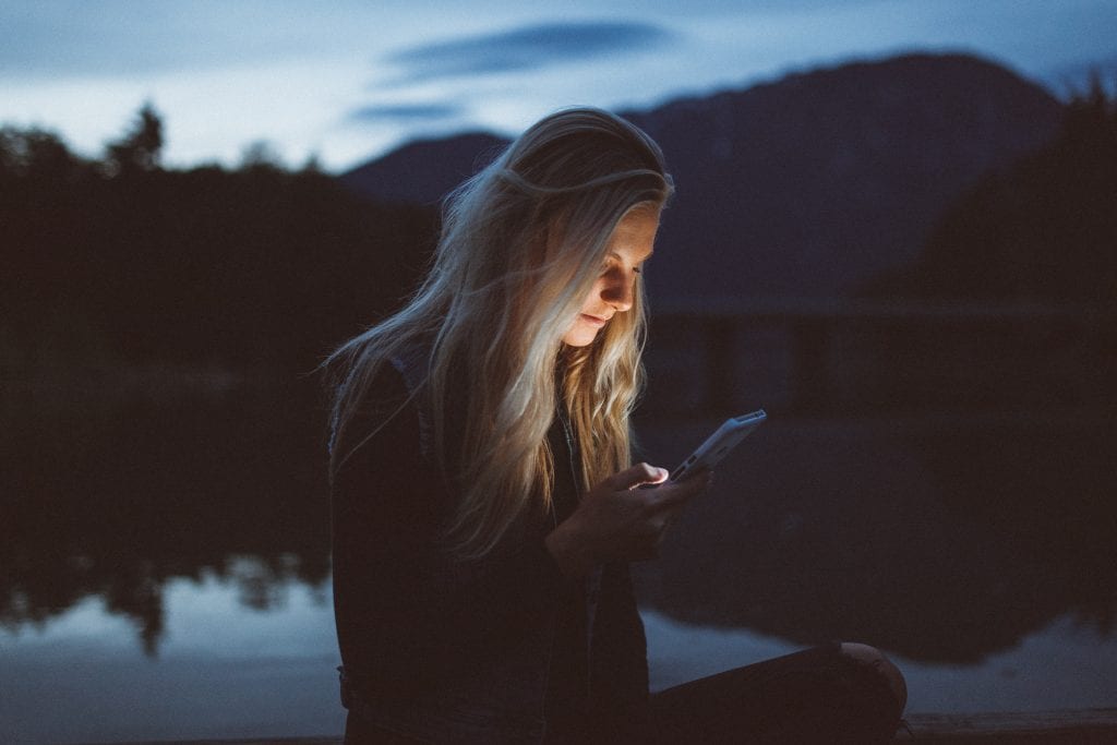 A woman standing near a body of water in the evening, looking at her cellphone. The light from the phone is illuminating her face.
