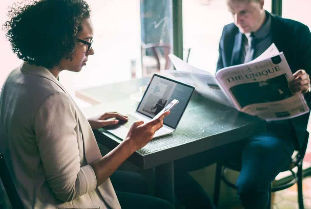 A women stops working at her laptop to check her mobile phone while sitting across from a business man reading a newspaper.