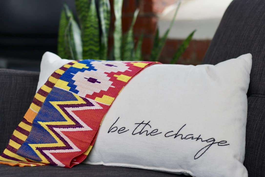 A Ghanian kente (tradition woven fabric) lays over a white pillow with "be the change" stitched in black along the bottom edge.