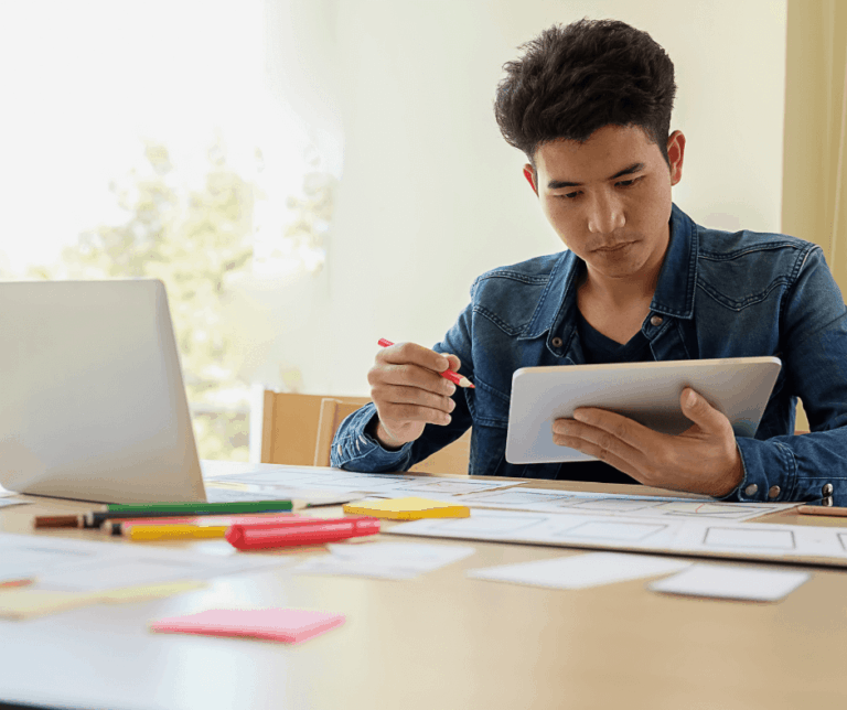 A man sits in front of a computer holding a notebook, post it notes and pens are around the desk representing our new for transitioning to and adopting digital technologies.