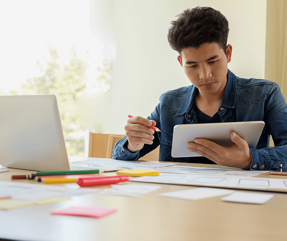 A man sits in front of a computer holding a notebook, post it notes and pens are around the desk