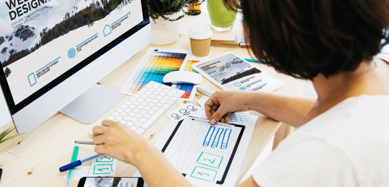 A woman with black hair sits at her desk with her computer in front of her. In her hands she is hand drawing website design features