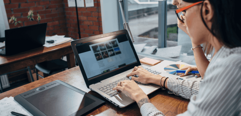 A woman with black hair wearing red glasses and a long sleeved shirt sits at a desk with her hands on the keyboard of a laptop. She is reviewing her website and a website care plan report describing the WordPress website maintenance performed.