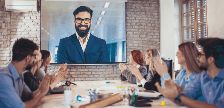 A variety of people are sitting around a boardroom table clapping a speaker on a large monitor