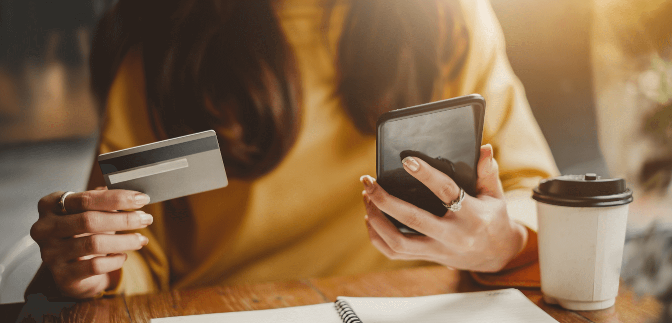 A woman in a yellow shirt holds her phone in her left hand and a credit card in her right hand. Her face is not showing.