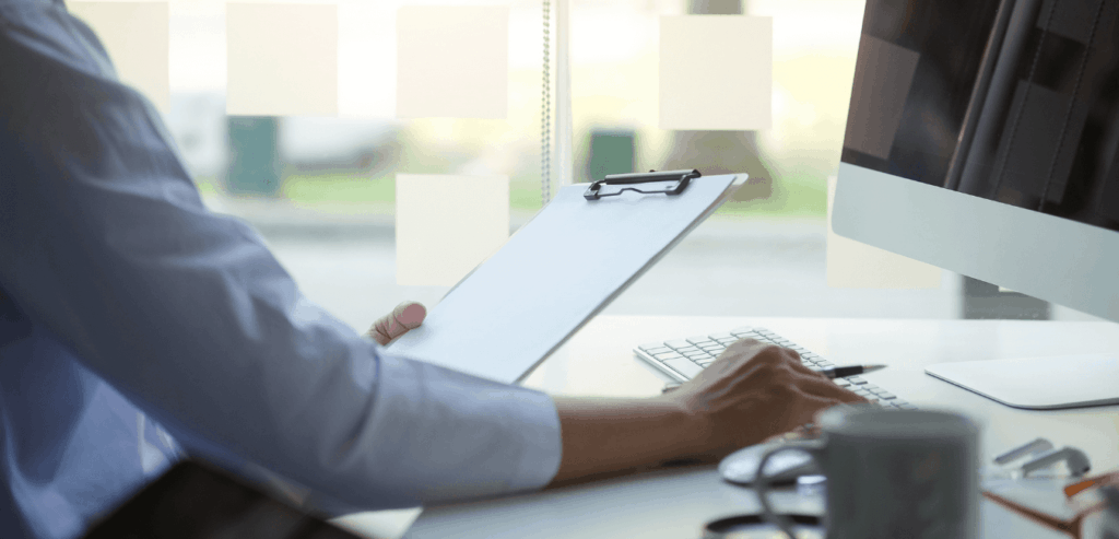 As seen in using donor data responsibly, a person sits at a desk, only their arm and part of their body is visible, the arm is outstretched to a computer and the other hand holds a clipboard.