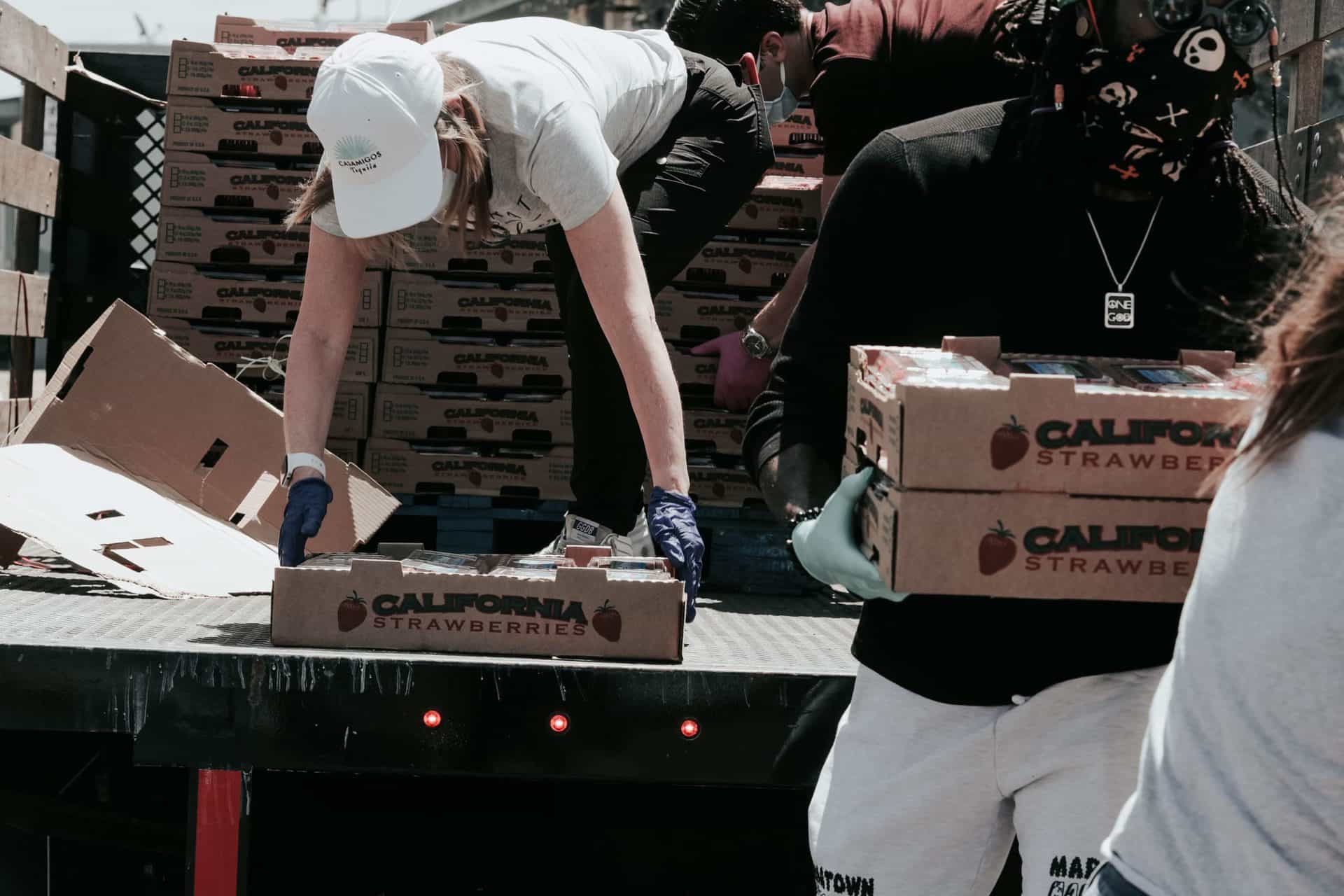 A woman bends down in the back of a truck picking up a box of food. She is wearing a white hat, white shirt and green pants. A man is walking away from the truck carrying boxes of food. It looks as if they are working at a food bank
