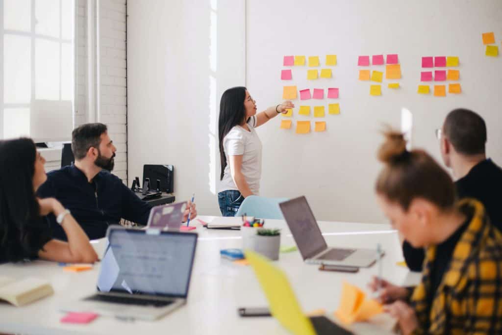 A woman stands at the front of a conference table pointing at post it notes on a wall. The conference table is surrounded by men and women with computers in front of them. Presumably they are all working together on a project