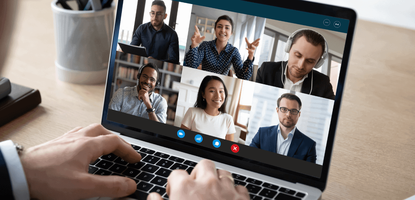 A man sits at a desk looking at a screen with 6 people in boxes on a conference call.