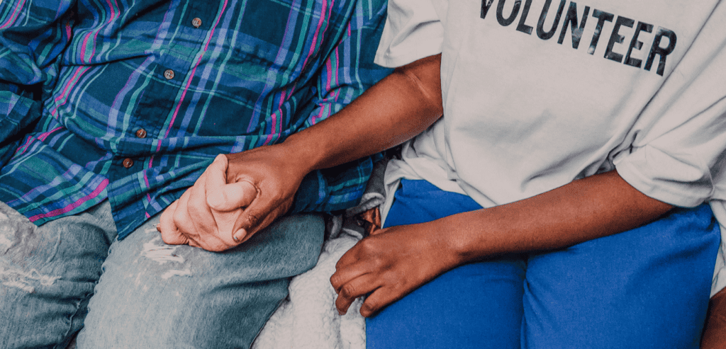 Two people sit next to each other holding hands. Only their torsos, arms and part of their legs are visible. The person on the right wears a shirt that says "volunteer"