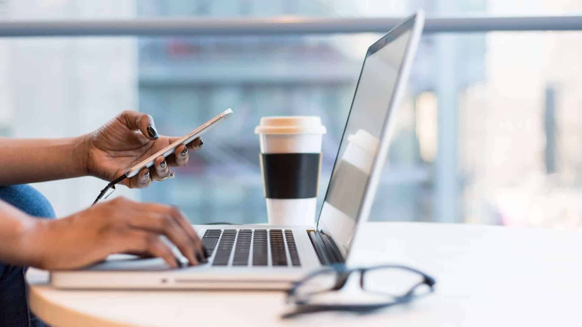 A woman's hands typing on a laptop while holding a smartphone.