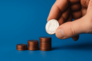 A hand places a dime on top of one of four ascending stacks of pennies arranged on a blue background.
