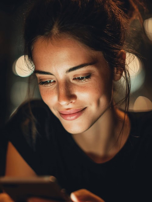 Young_Woman_Viewing_a_Website_on_Her_Smartphone A woman with dark hair smiles softly while looking at her smartphone in low, warm lighting.