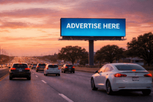 Cars drive on a highway at sunset with a large digital billboard above displaying the message “ADVERTISE HERE.”.