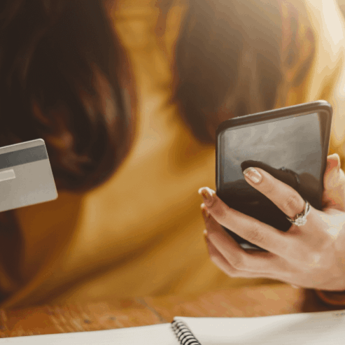 A woman in a yellow shirt holds her phone in her left hand and a credit card in her right hand. Her face is not showing.