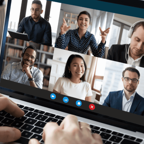 A man sits at a desk looking at a screen with 6 people in boxes on a conference call.