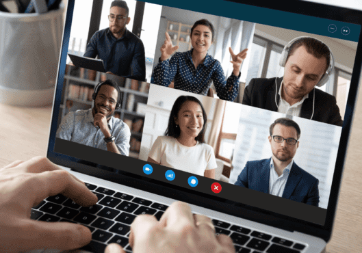 A man sits at a desk looking at a screen with 6 people in boxes on a conference call.