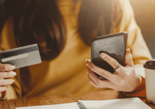 A woman in a yellow shirt holds her phone in her left hand and a credit card in her right hand. Her face is not showing.