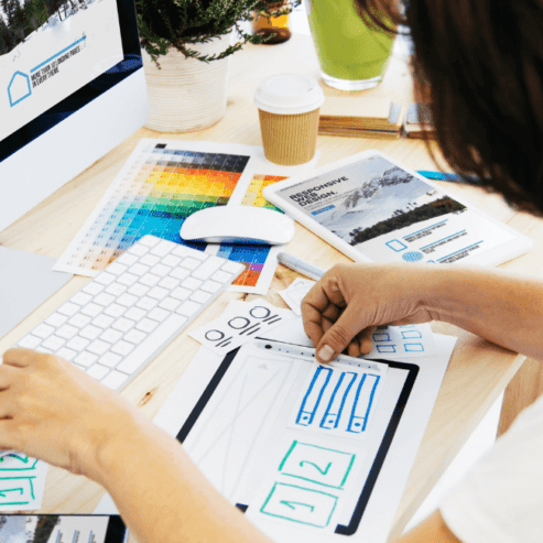 A woman with black hair sits at her desk with her computer in front of her. In her hands she is hand drawing website design features