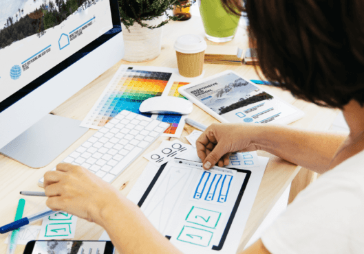 A woman with black hair sits at her desk with her computer in front of her. In her hands she is hand drawing website design features