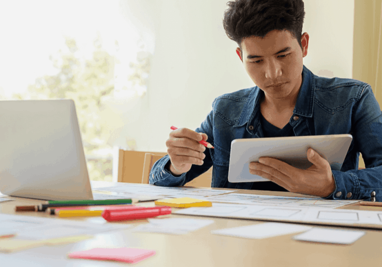 A man sits in front of a computer holding a notebook, post it notes and pens are around the desk representing our new for transitioning to and adopting digital technologies.