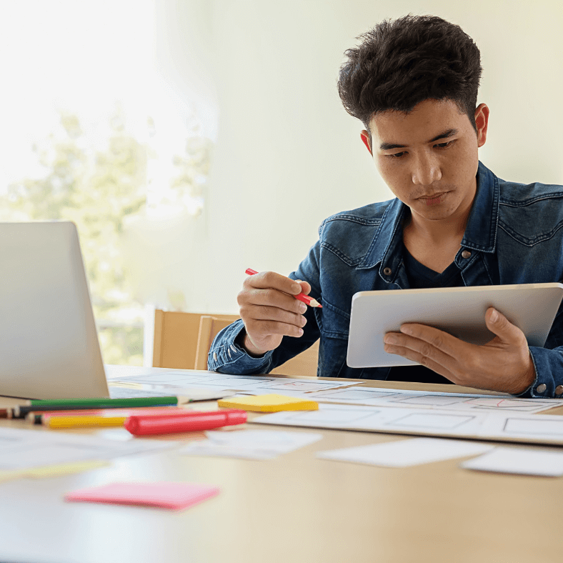 A man sits in front of a computer holding a notebook, post it notes and pens are around the desk