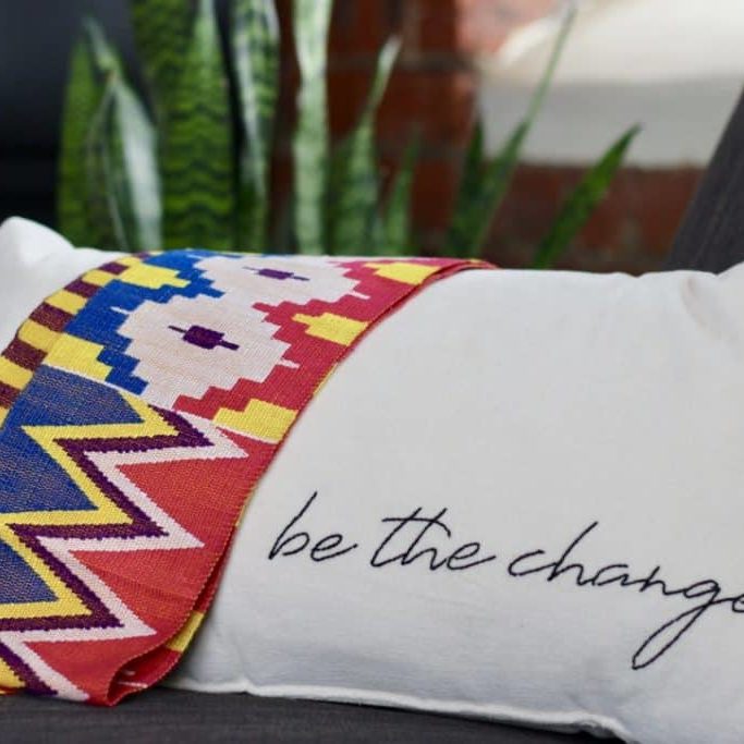 A Ghanian kente (tradition woven fabric) lays over a white pillow with "be the change" stitched in black along the bottom edge.