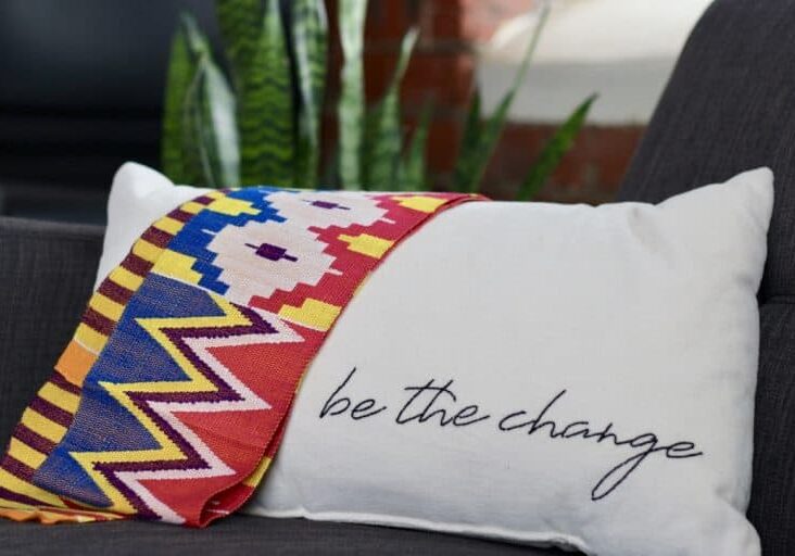 A Ghanian kente (tradition woven fabric) lays over a white pillow with "be the change" stitched in black along the bottom edge.