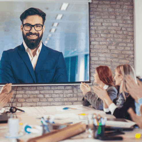 A variety of people are sitting around a boardroom table clapping a speaker on a large monitor