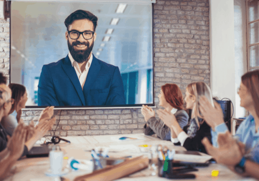 A variety of people are sitting around a boardroom table clapping a speaker on a large monitor