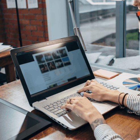 A white woman wearing red glasses and a long sleeved shirts sits at a table with her hands on the keyboard of a laptop. She is reviewing a website care plan report describing the WordPress website maintenance performed.