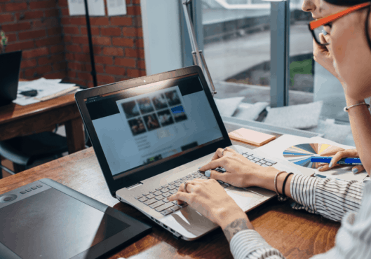 A woman with black hair wearing red glasses and a long sleeved shirt sits at a desk with her hands on the keyboard of a laptop. She is reviewing her website and a website care plan report describing the WordPress website maintenance performed.