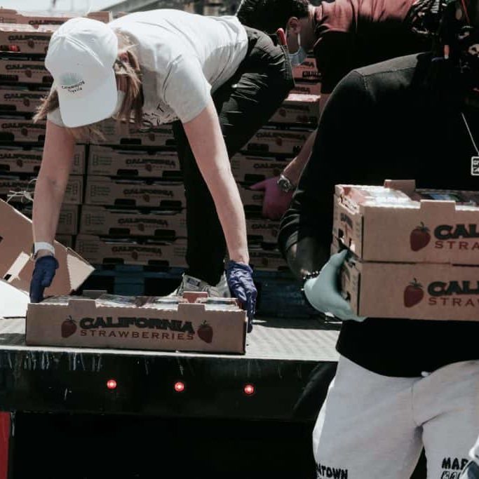 A woman bends down in the back of a truck picking up a box of food. She is wearing a white hat, white shirt and green pants. A man is walking away from the truck carrying boxes of food. It looks as if they are working at a food bank