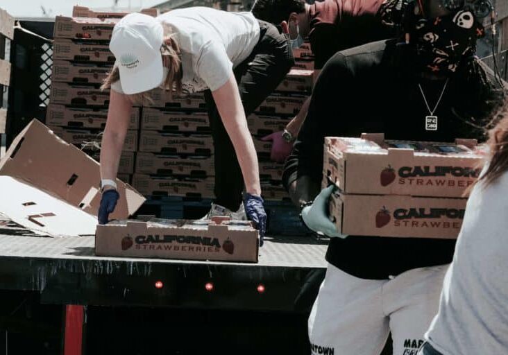 A woman bends down in the back of a truck picking up a box of food. She is wearing a white hat, white shirt and green pants. A man is walking away from the truck carrying boxes of food. It looks as if they are working at a food bank