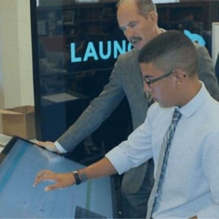 A student interacts with the Launch to Learn pltform using a large touch screen monitor while being filmed by news media.
