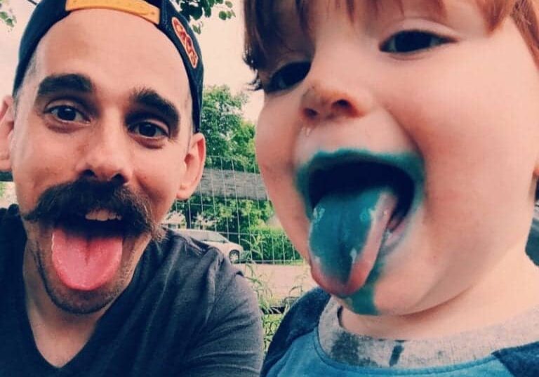 A man and child stick their tongues out. The child's tongue is blue from candy